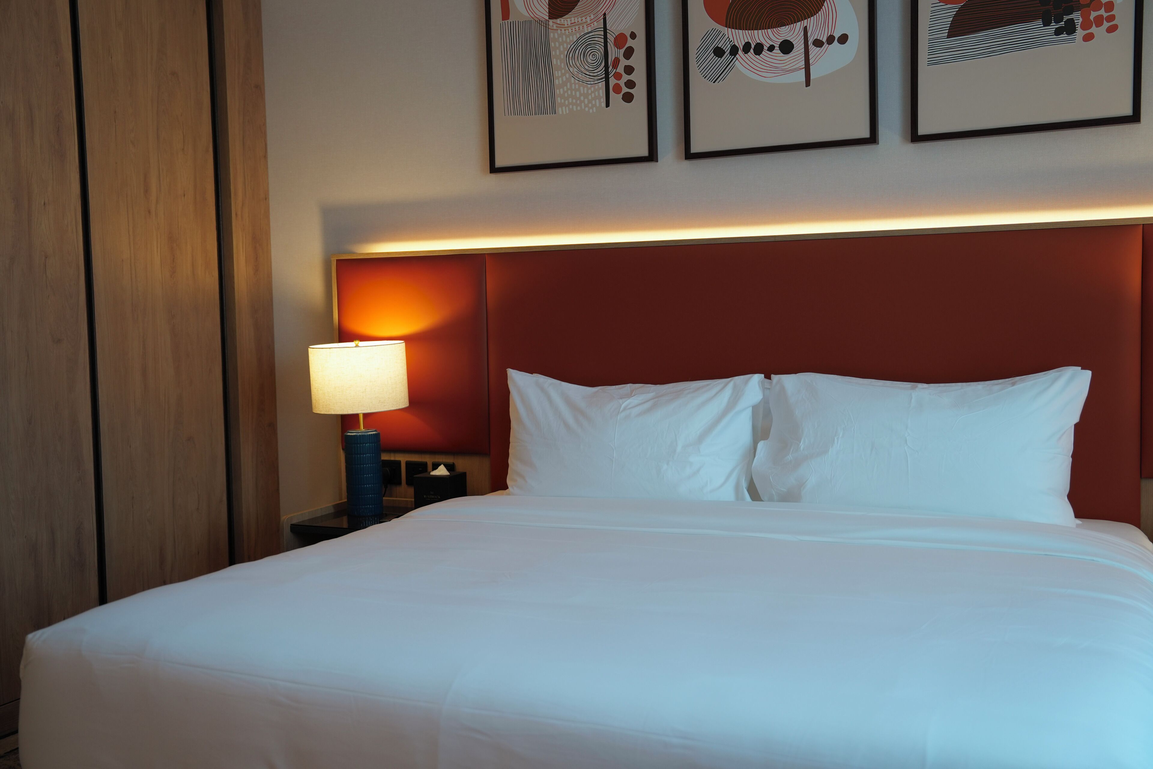 Een hotelkamer met een groot bed, een nachtkastje met een lamp, en drie ingelijste kunstwerken aan de muur.