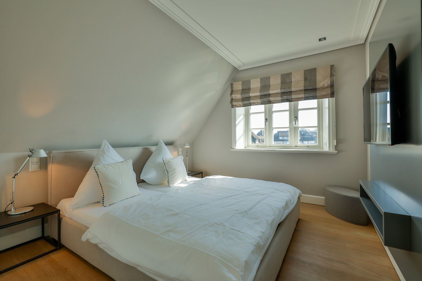 A bedroom with a bed, bedside lamps, a window with curtains, a flat-screen TV, and a small bedside table.