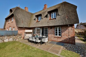 A traditional house with a thatched roof, a stone wall, and a patio with a table and chairs. - Sand Beach - Luxury vacation home Sand Beach in Rantum - with sauna, 450 meters from the beach (Sylt)