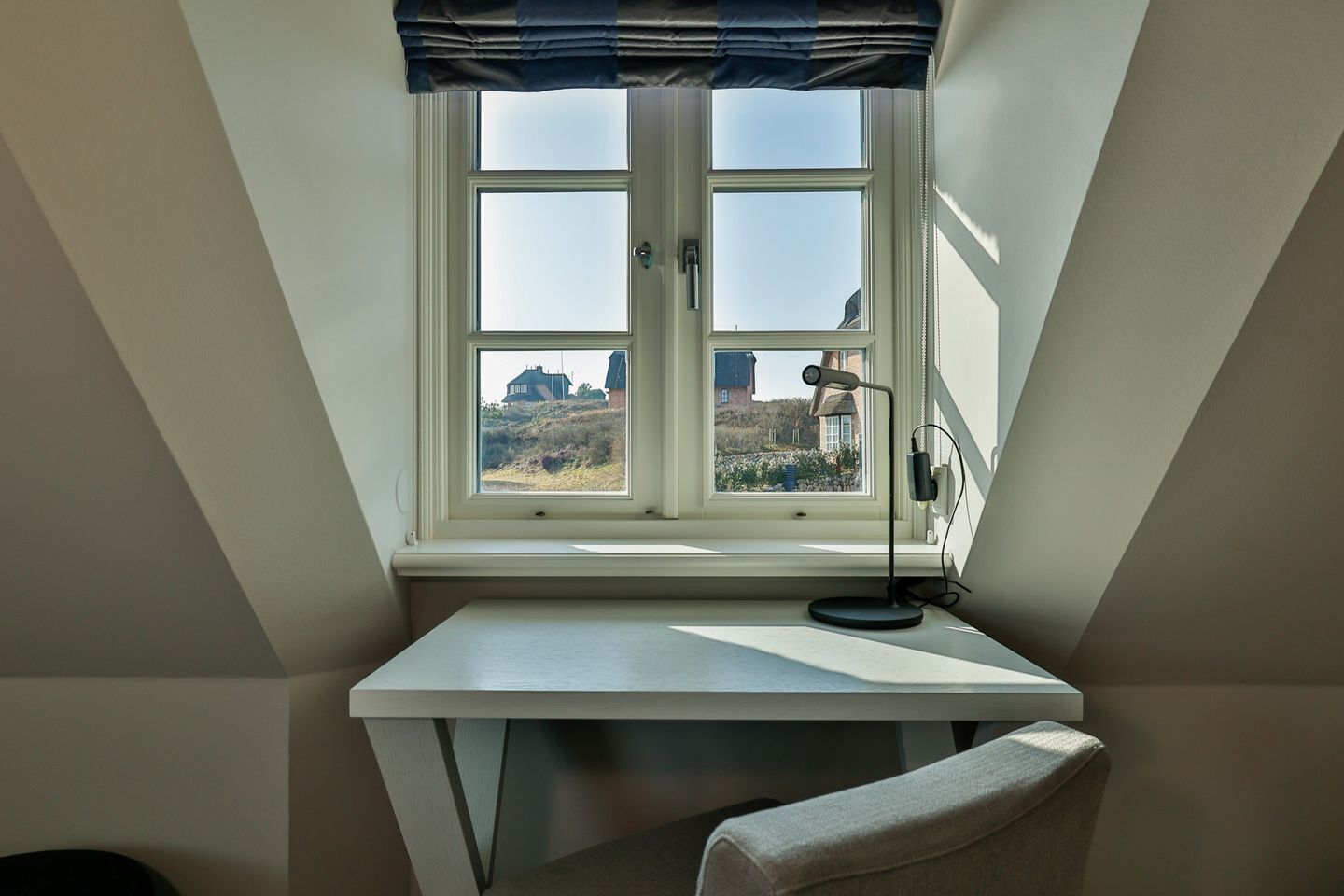 A room with a desk, a chair, a window with a view of buildings, and a lamp.