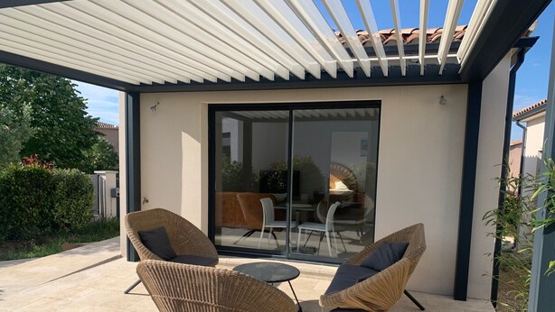 En täckt uteplats med två fåtöljer i rotting och ett litet bord, en skjutdörr i glas som leder ut till en utomhusplats, och en pergola ovanför.