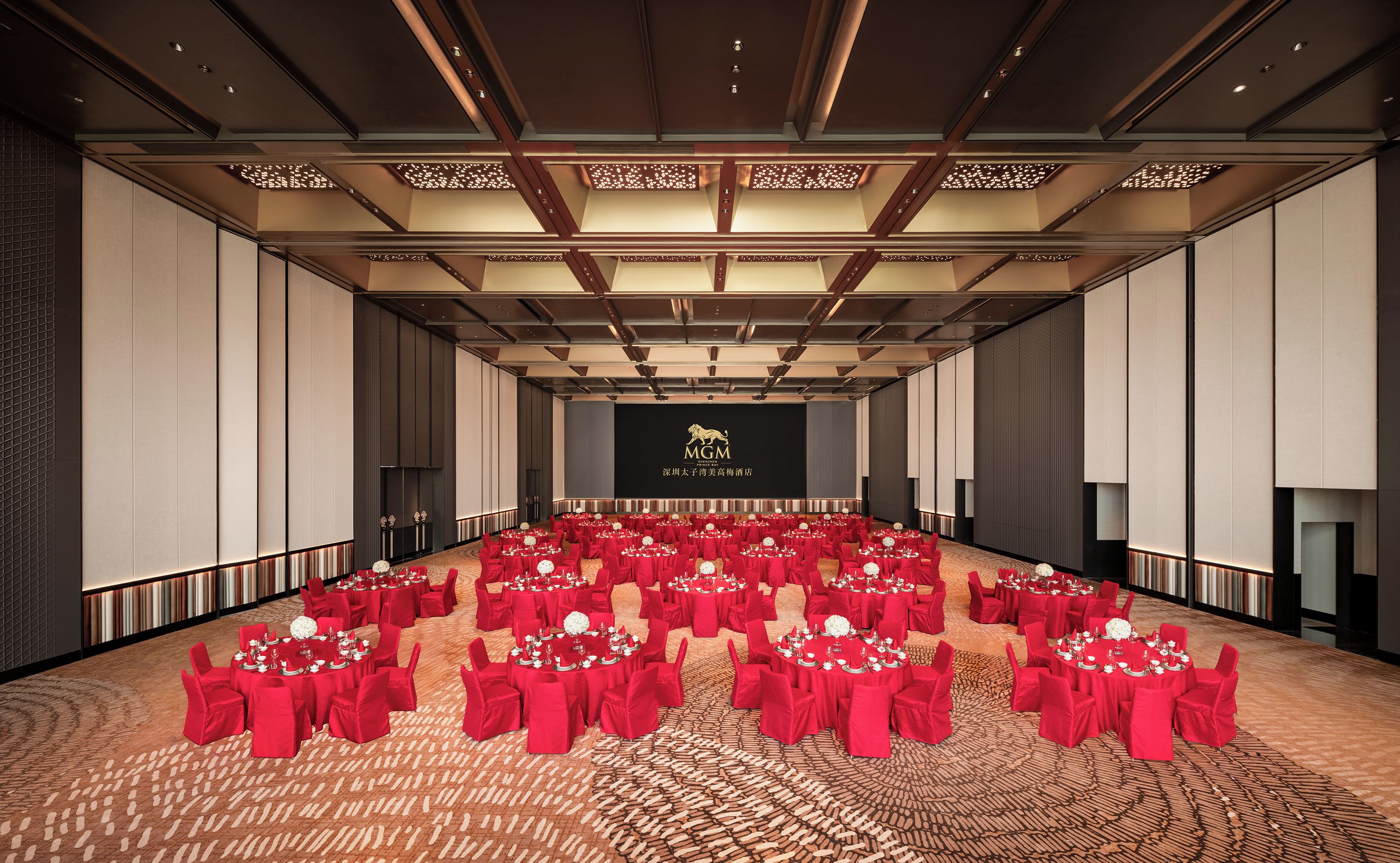 A large conference room with rows of tables and chairs, a stage with a logo, and a patterned carpet.