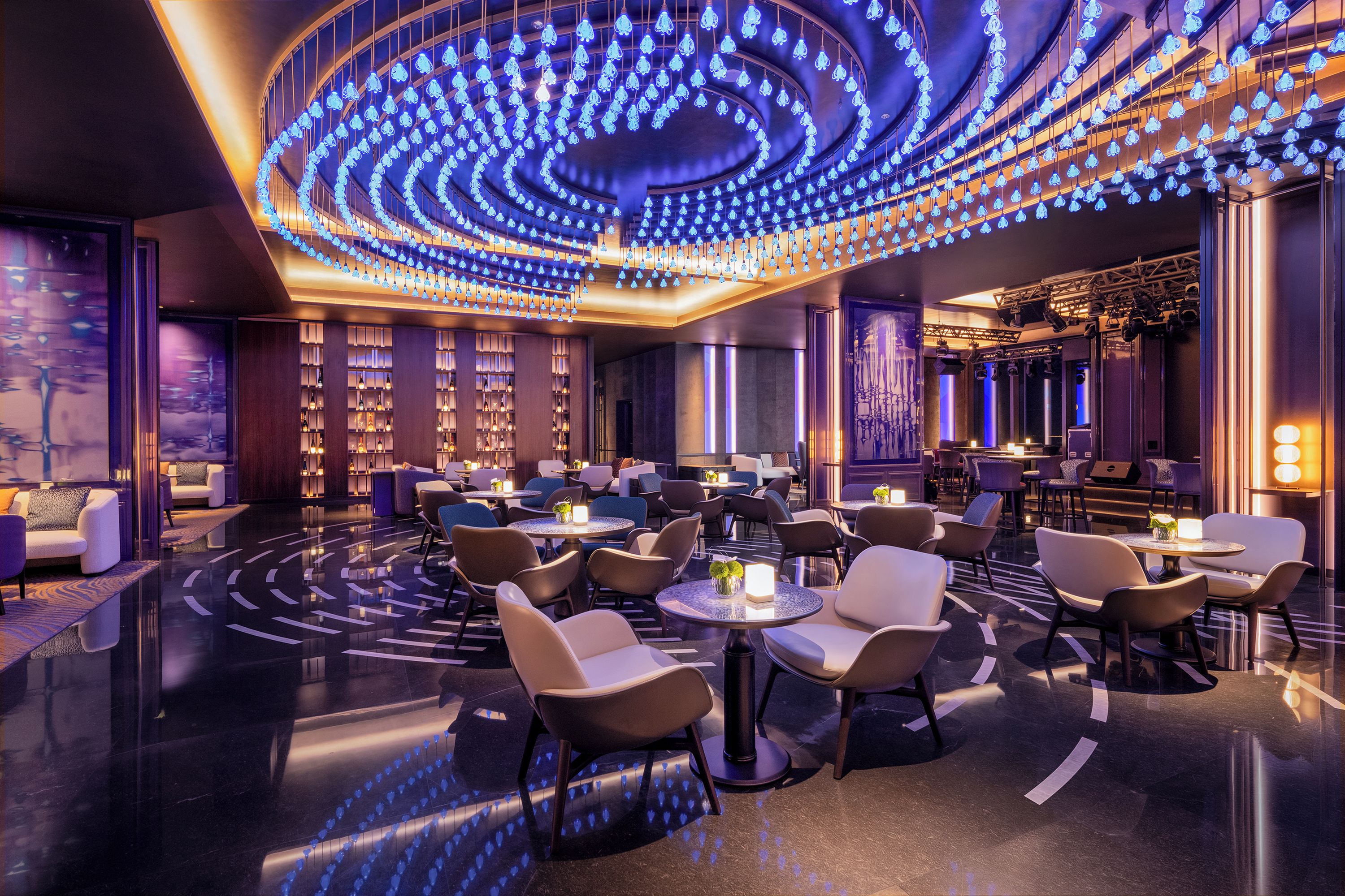 A modern hotel lobby with a circular ceiling light installation, seating areas, and a bar.