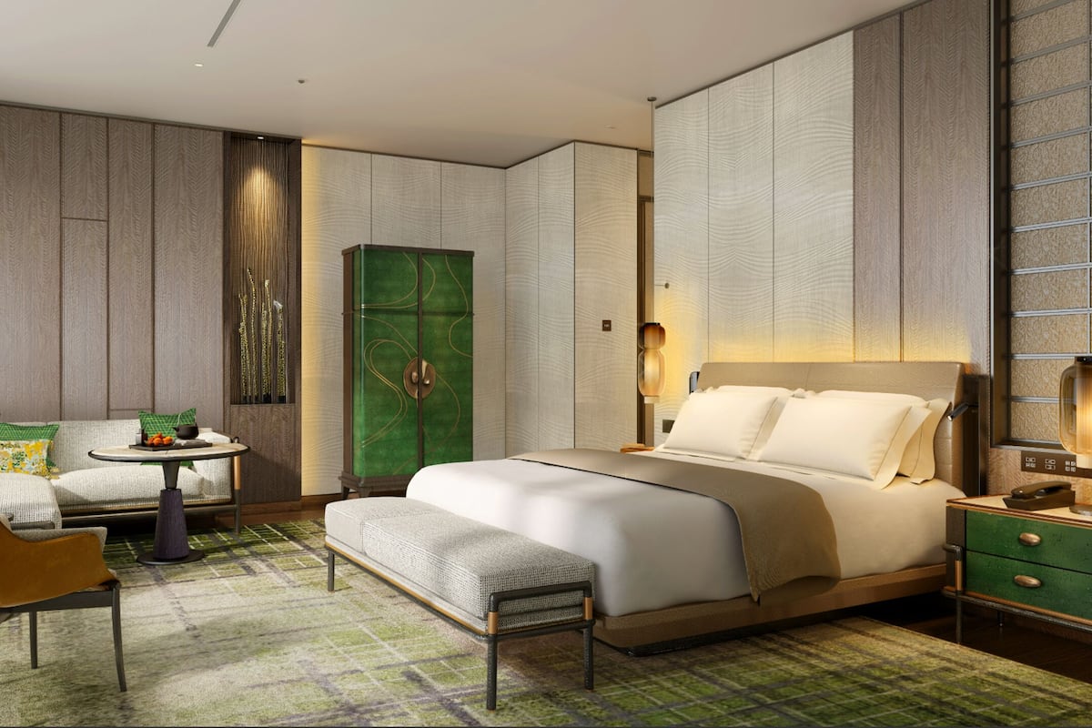 A hotel room with a bed, a seating area with a table and chairs, a green cabinet, and a nightstand with a lamp.
