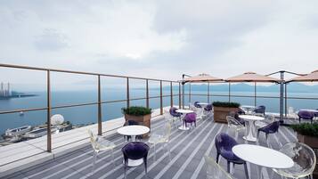 A rooftop terrace with clear views of the sea and city skyline, featuring modern seating and potted plants.