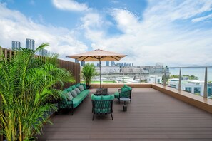 Une terrasse sur le toit, aménagée avec un canapé, des chaises et un parasol, d’où l’on peut admirer une vue panoramique sur la ville et l’étendue d’eau.