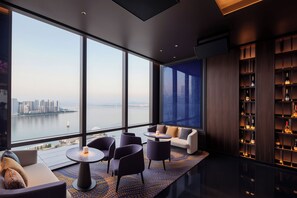 Een modern interieur met een groot raam dat uitkijkt op een stadsbeeld, een zithoek met een ronde tafel en een wijnrek.