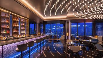 A modern bar area with a city view, bar stools, and a variety of bottles on shelves.