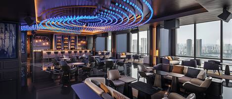 A modern lounge with a curved ceiling light fixture, seating arrangements, and a city view.