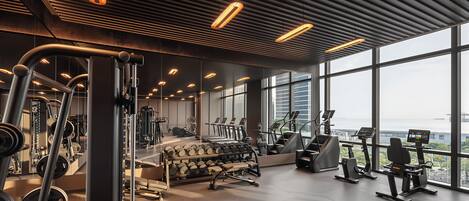 A modern gym with various exercise equipment, large windows, and a view of the city.