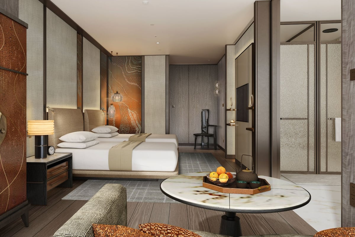 A modern hotel room with a bed, bedside table, lamp, and a round table with fruit.
