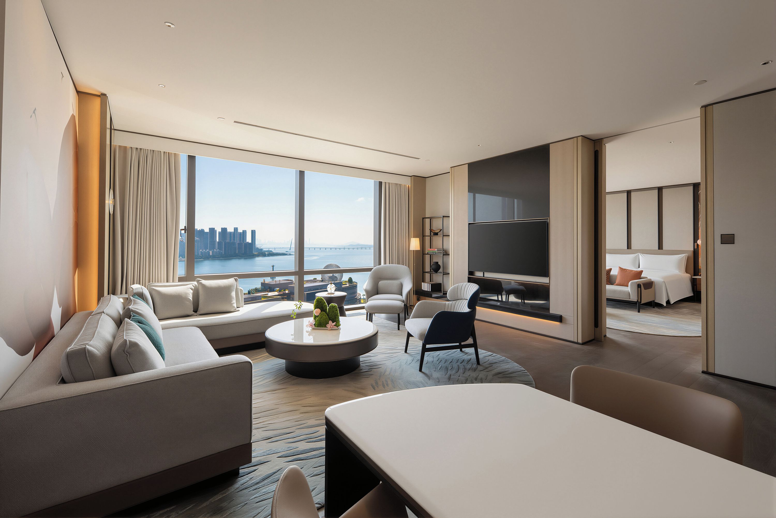 A modern hotel room with a large window offering a city view, a sofa, a dining table, and a separate seating area.