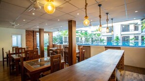 Un restaurante con mesas y sillas de madera, lámparas colgantes y vistas a edificios y zonas verdes al exterior.