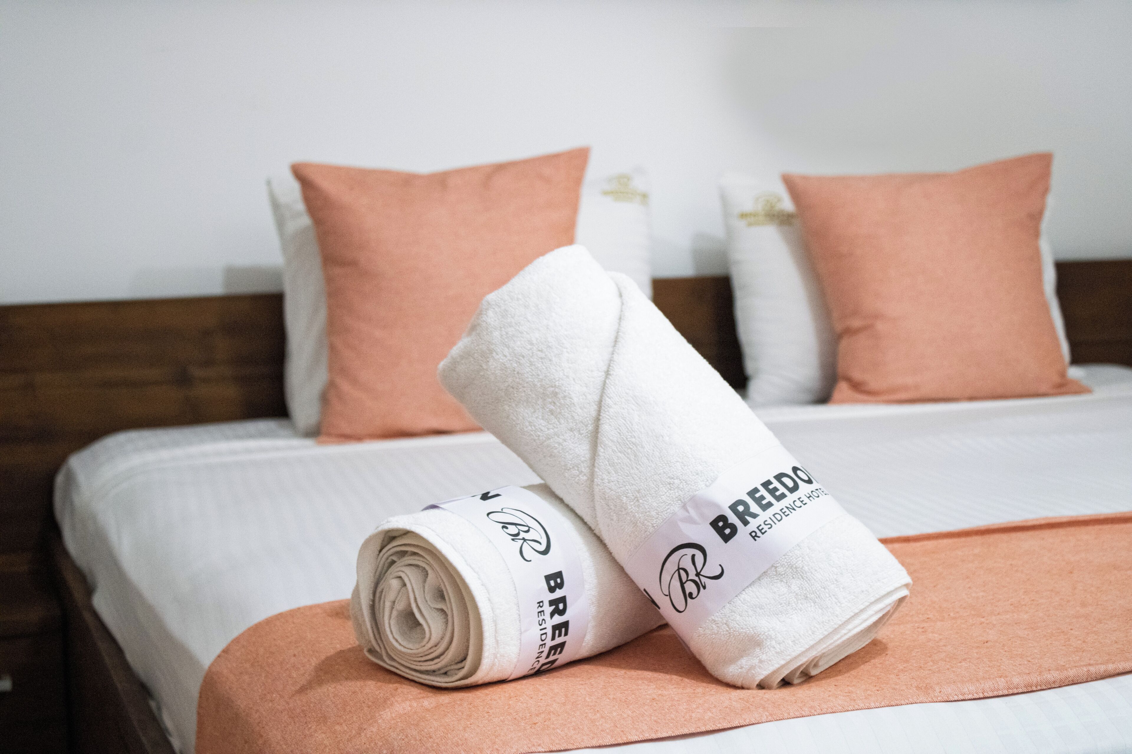 Una cama bien tendida con una toalla enrollada y una almohada, un cabecero de madera y una pared blanca.