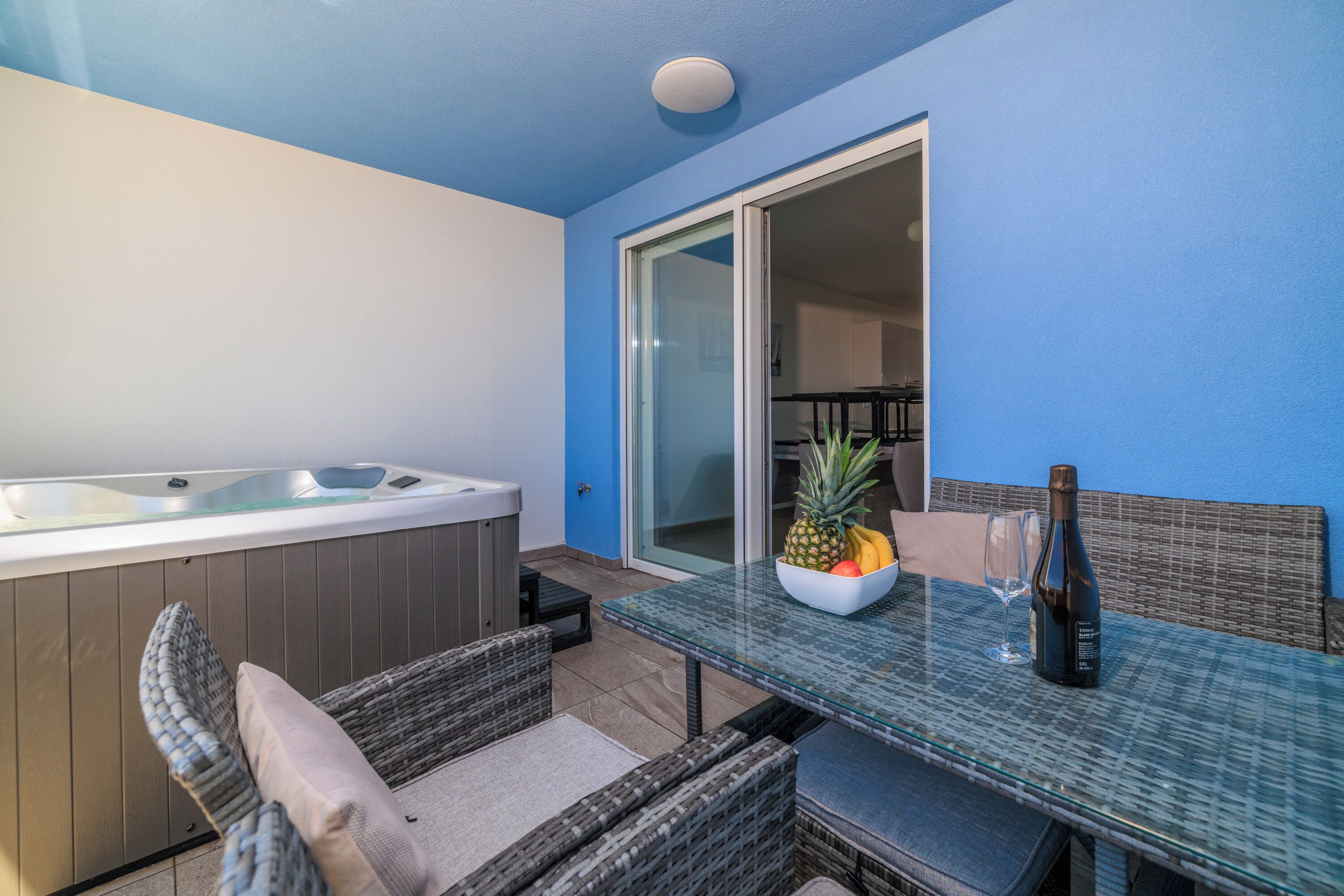Ein Balkon mit Whirlpool, Korbmöbeln, einem Glastisch mit Obstschale und Weinflasche sowie einer Schiebetür zu einem Innenbereich.