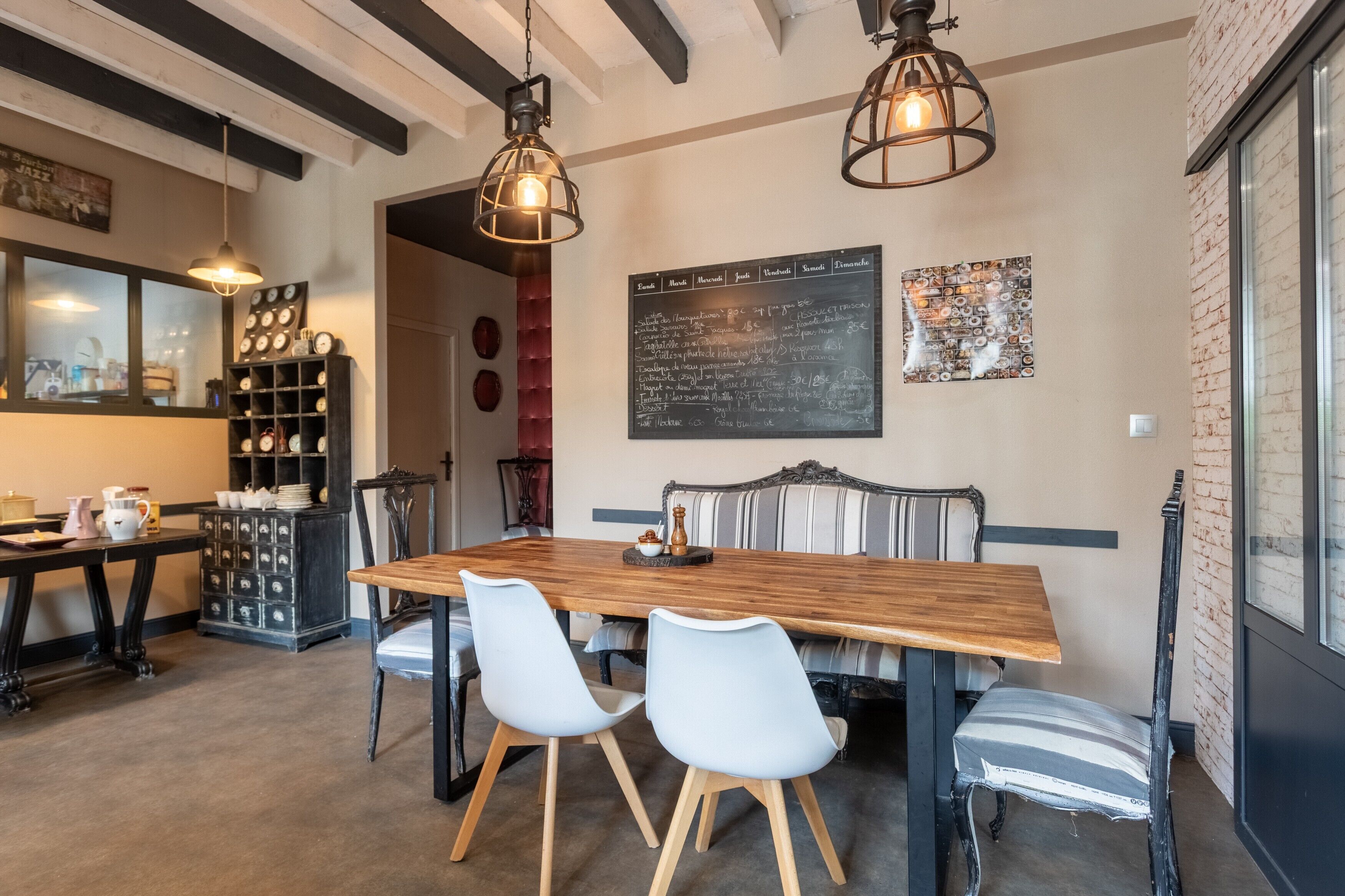 Un espace repas avec une table en bois, quatre chaises, deux luminaires suspendus, un tableau noir servant de menu et un mur en briques.