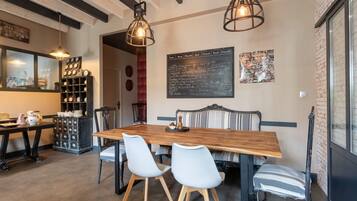 Un espace repas avec une table en bois, quatre chaises, deux luminaires suspendus, un tableau noir servant de menu et un mur en briques.