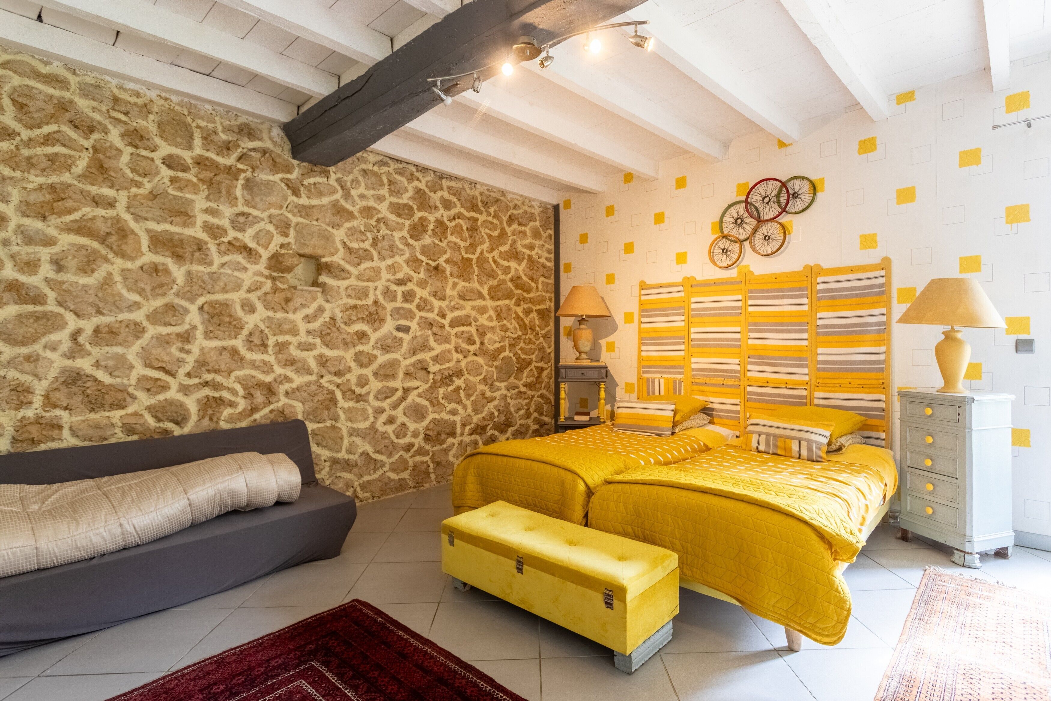Un dormitorio con pared de piedra, cama con ropa de cama amarilla, sofá gris y banco de madera.
