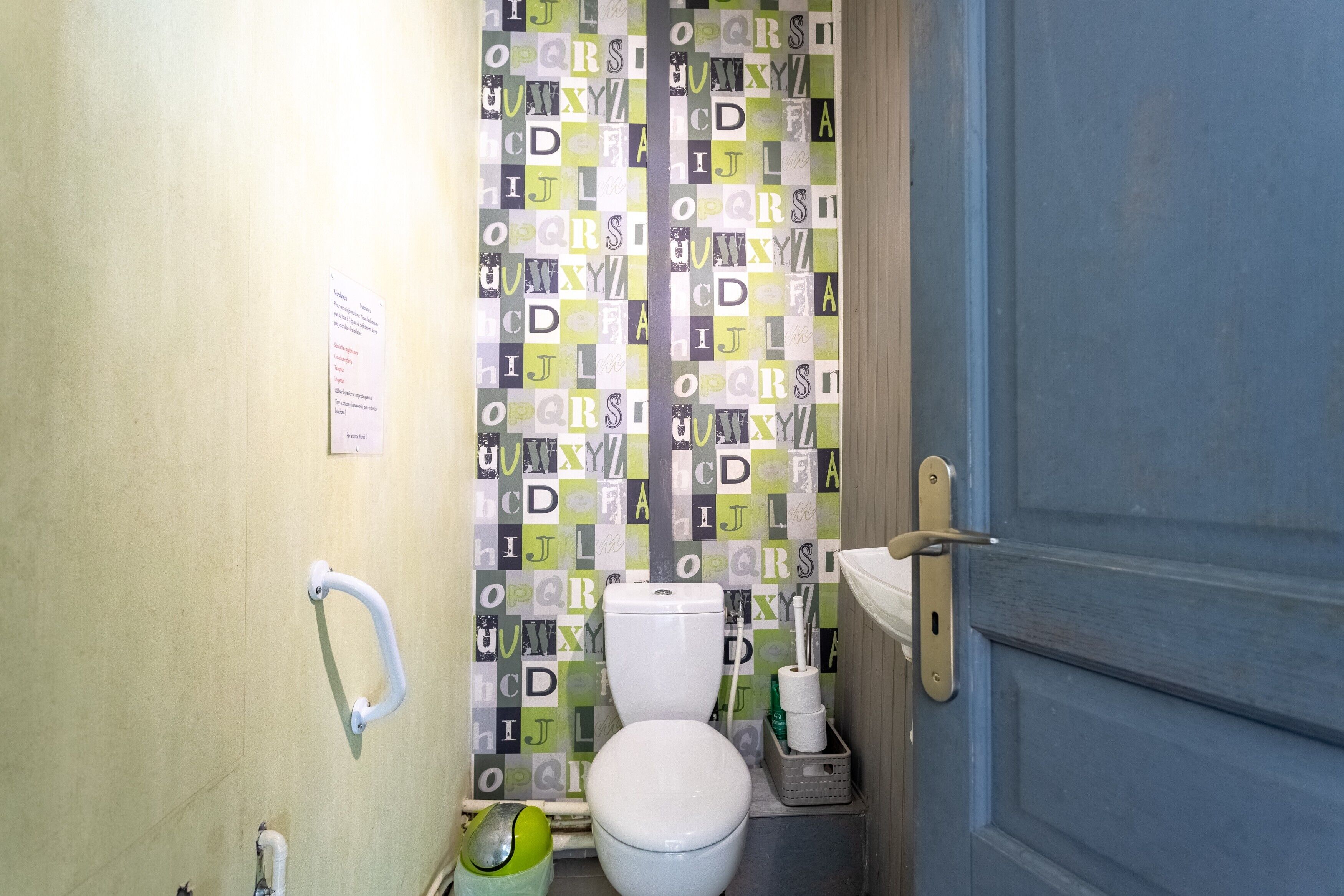 A small bathroom with a toilet, a handrail, and a shower curtain with alphabet patterns.