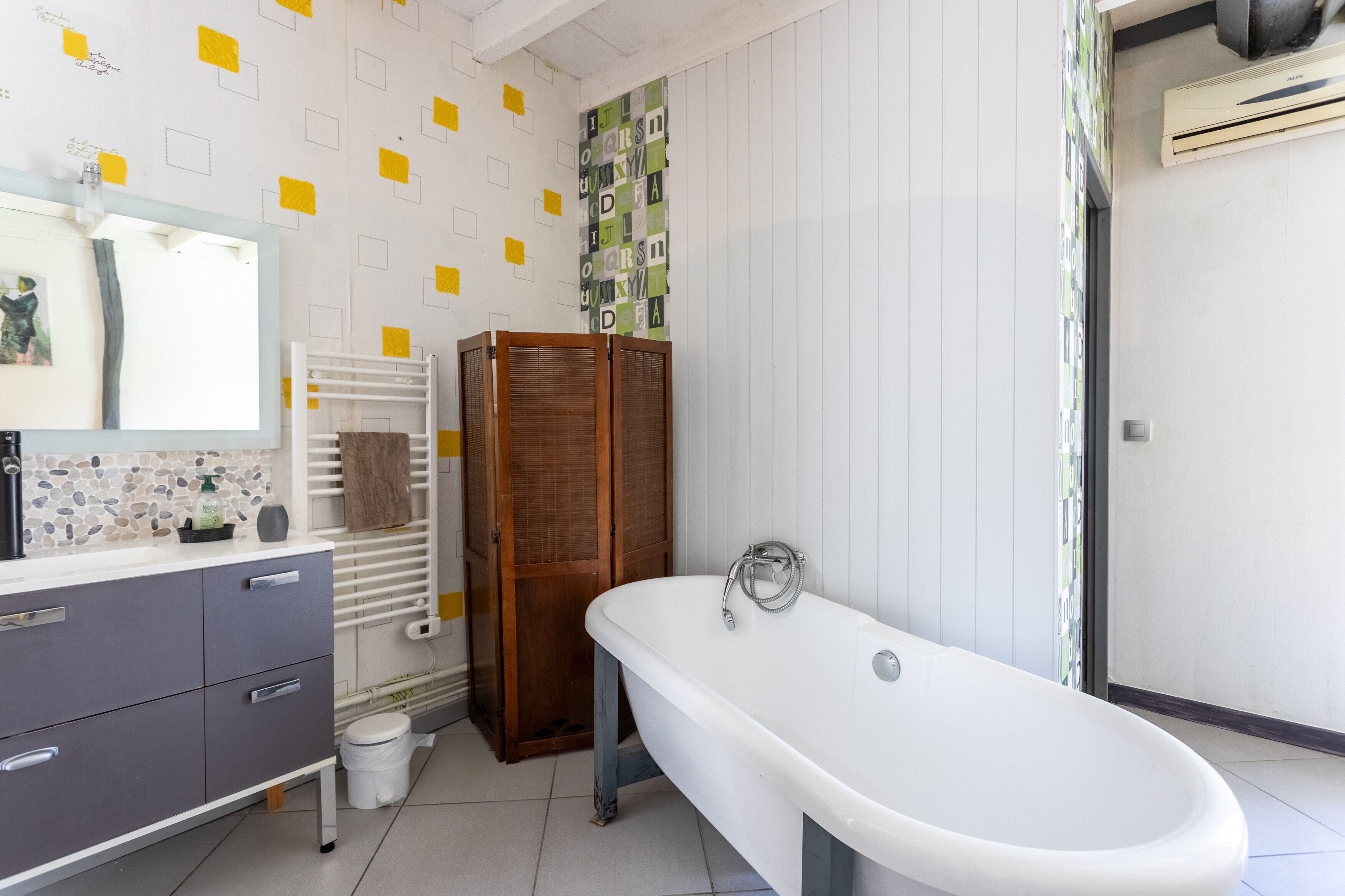 Ein Badezimmer mit einer weißen Badewanne, einem Waschtisch mit Schubladen, einem Spiegel, einem Handtuchhalter und einer Wand mit geometrischem Muster.