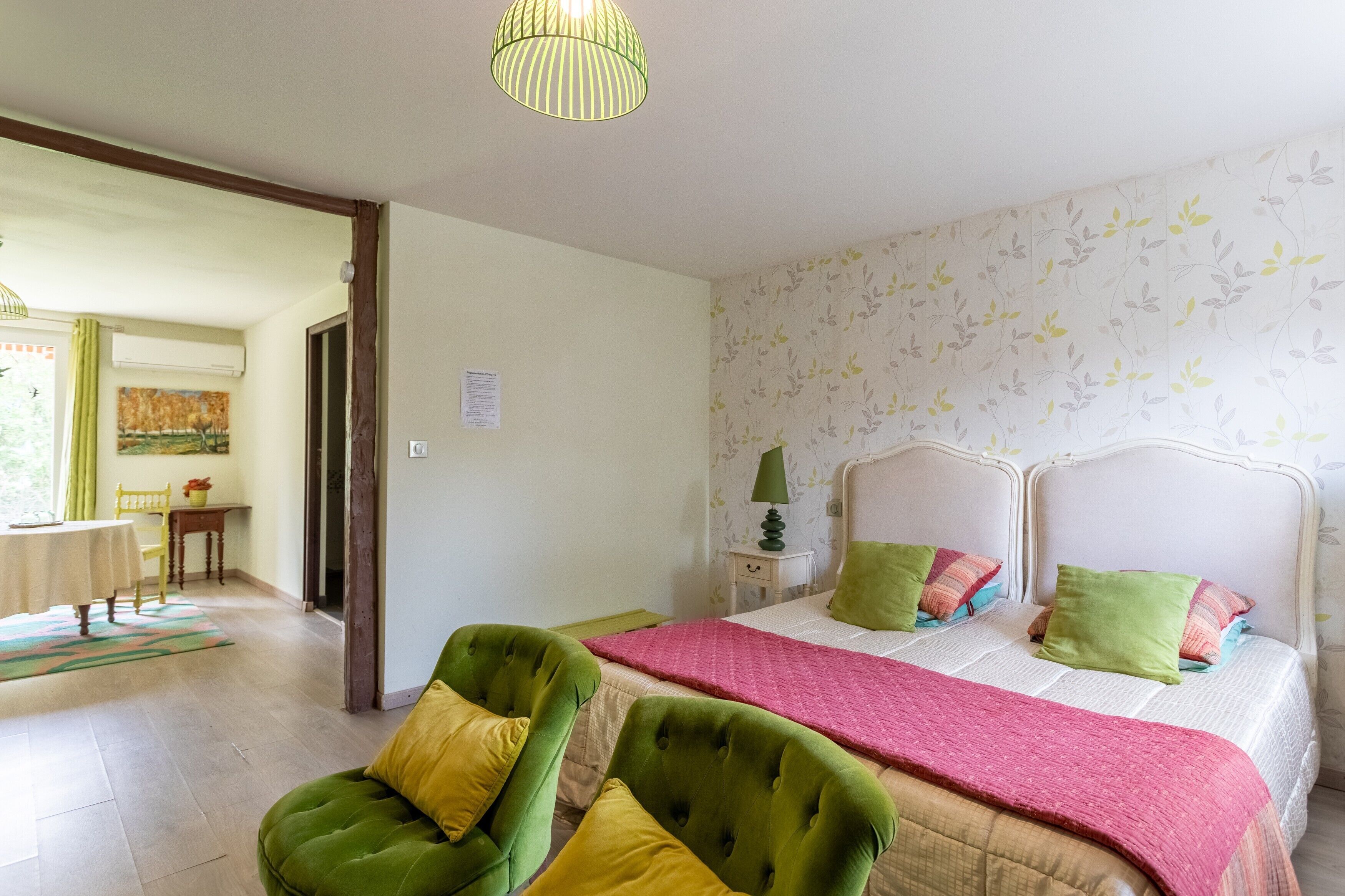 Une chambre à coucher avec un lit, des chaises vertes et jaunes, une table de chevet, une lampe et une fenêtre avec des rideaux.
