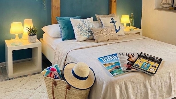 Een slaapkamer met schuine plafonds, een bed met veel kussens, nachtlampjes, een kleine plant en een tijdschrift op het bed.