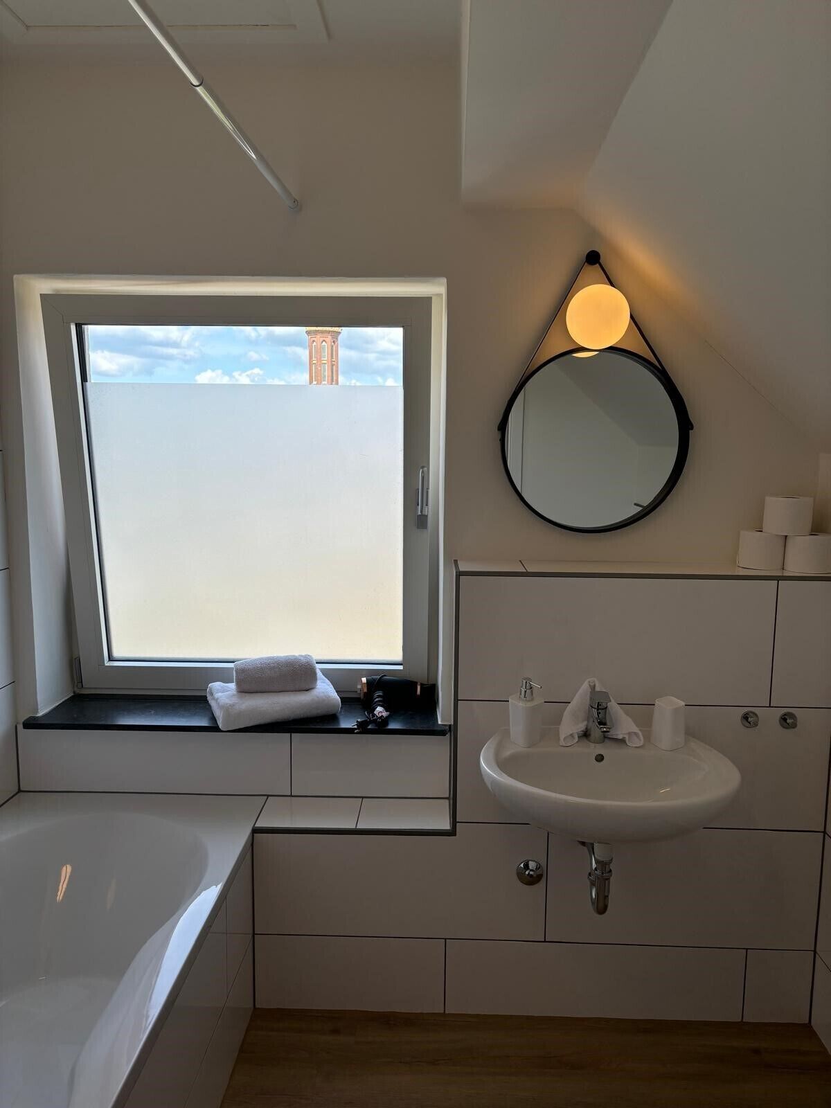 Un baño moderno con ventana, espejo redondo, lavamanos y bañera.