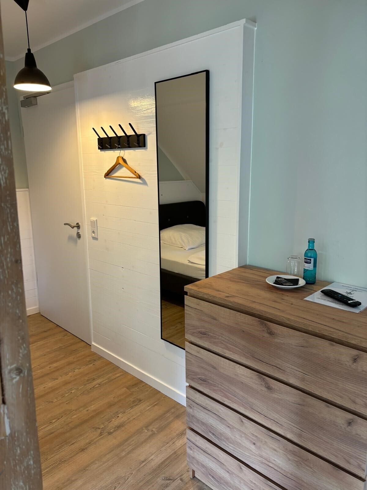 A bedroom with a wooden dresser, a hanging coat rack, a bed, and a mirror.