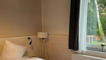 A hotel room with a bed, bedside lamp, window with curtains, and a view of trees outside.
