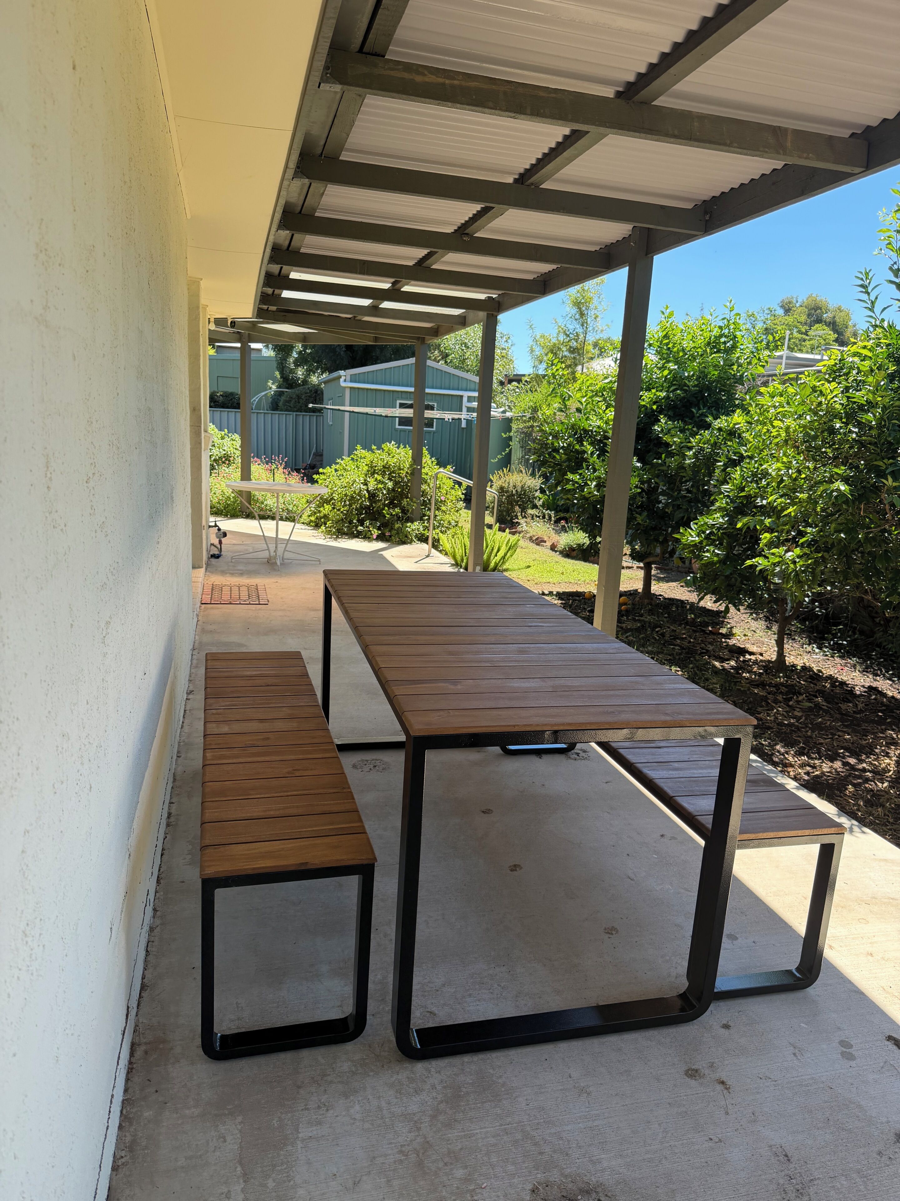 Um pátio externo com área coberta, dois bancos de madeira e uma mesa de metal.