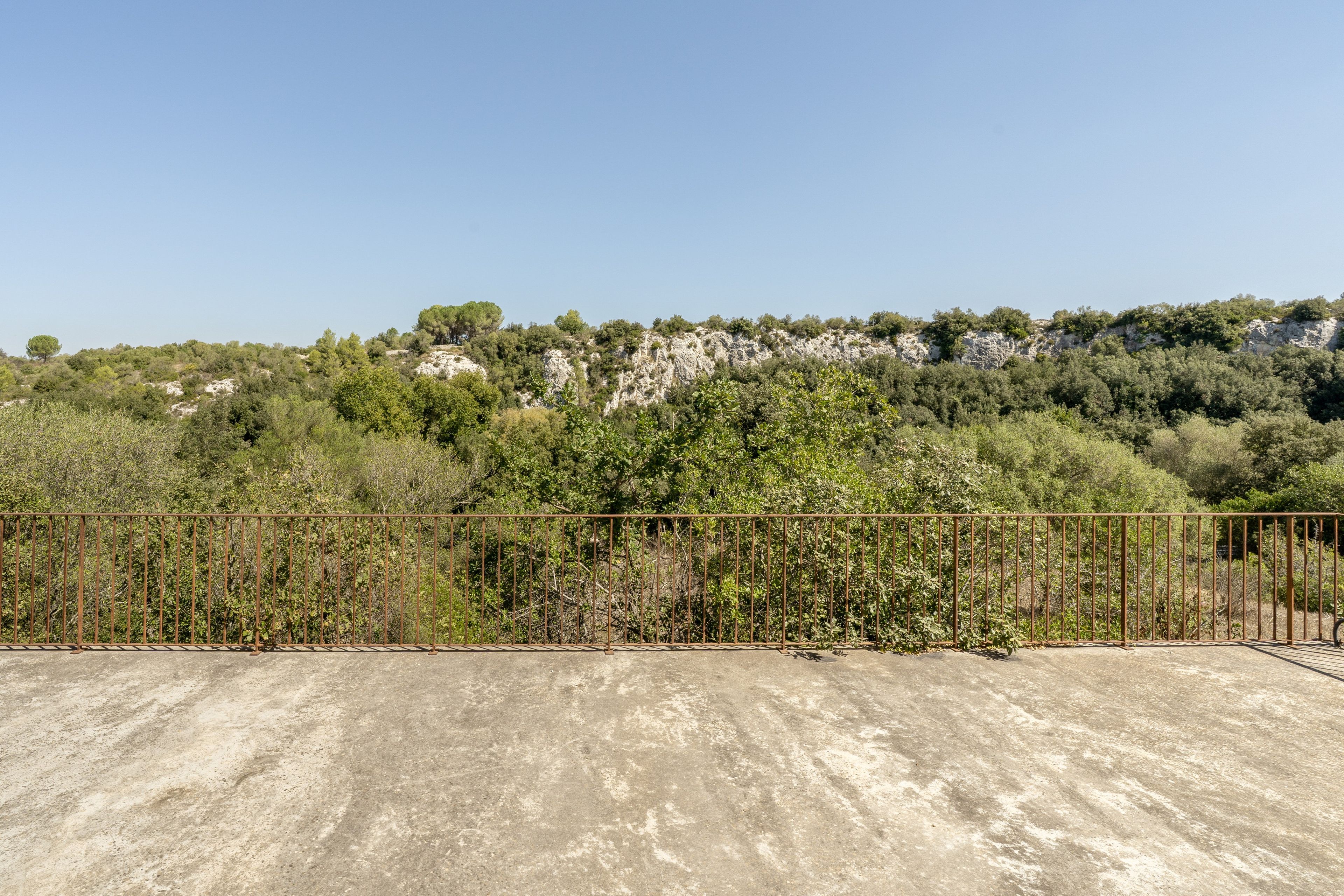 Eine Betonfläche mit einem Metallzaun im Vordergrund, ein klarer Himmel und ein Hügel mit Bäumen und Felsformationen im Hintergrund.