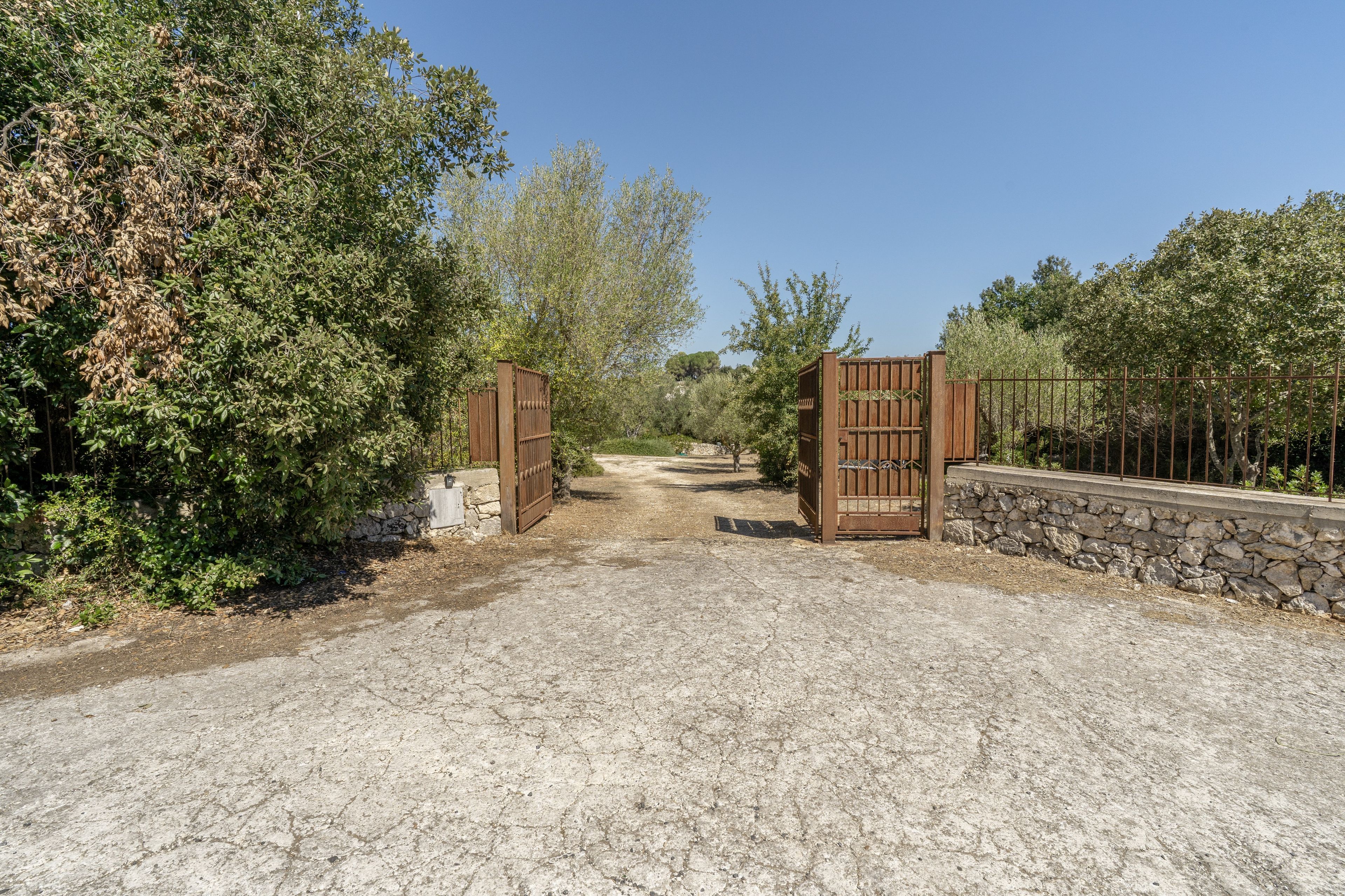 A gravel driveway leading to a gated entrance with a stone wall on one side and dense foliage on the other.