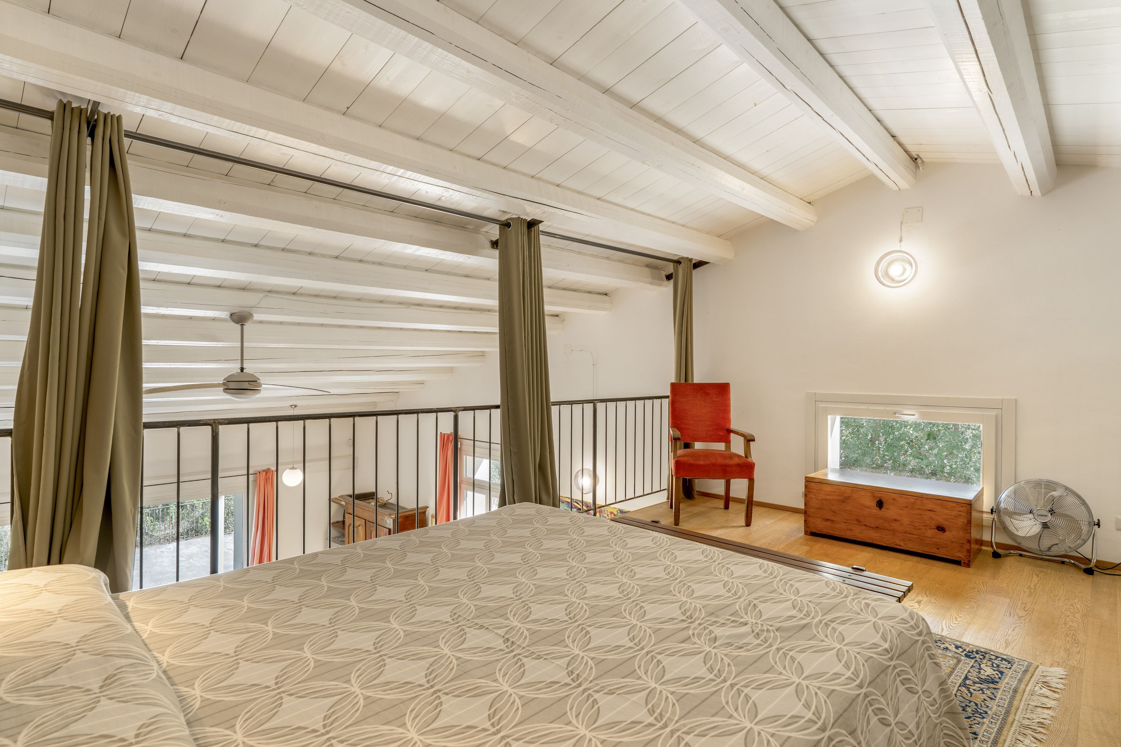 A bedroom with a large bed, a red chair, a wooden dresser, a ceiling fan, and a window with a view.