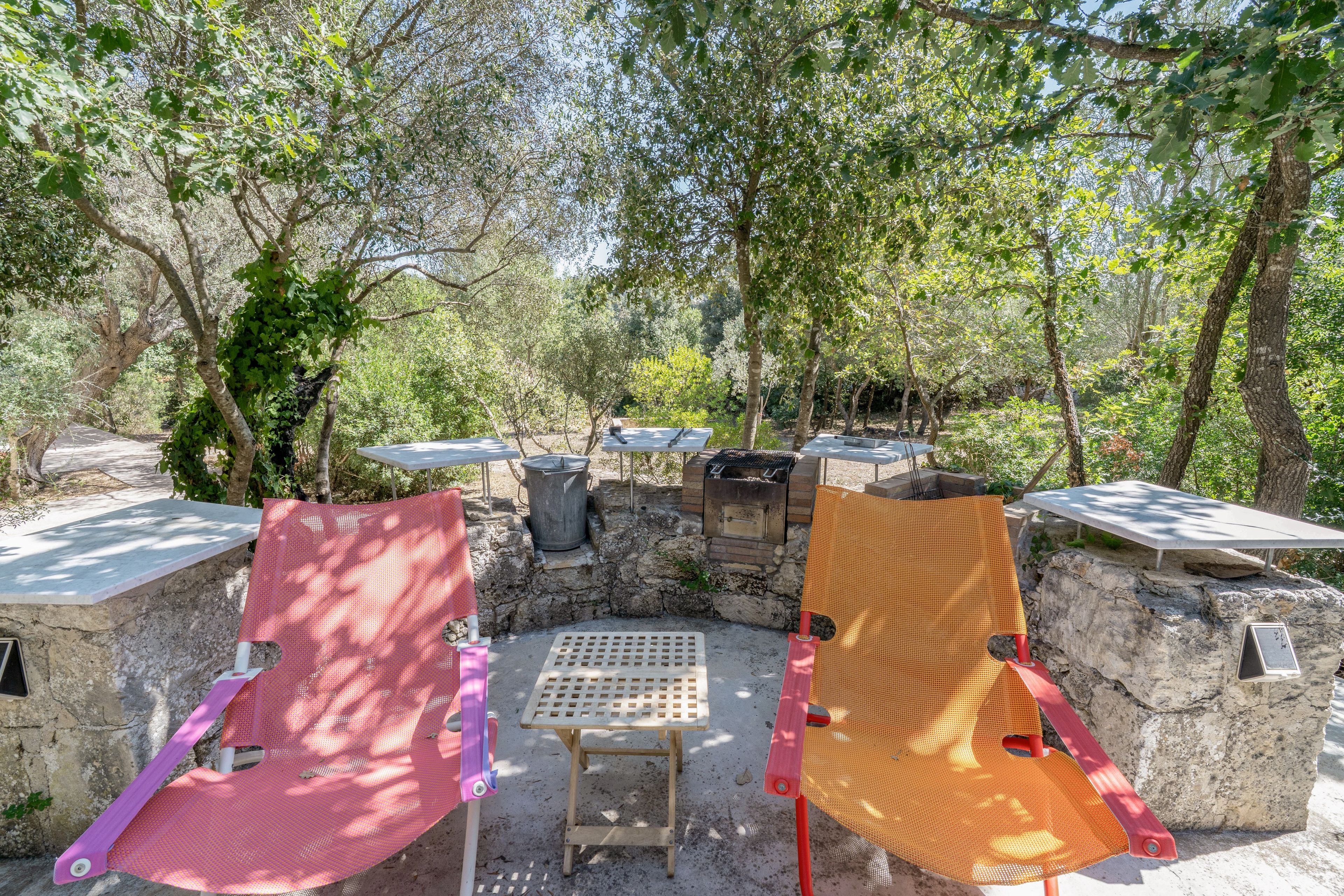 An outdoor seating area with two chairs and a table, surrounded by trees and a stone wall.