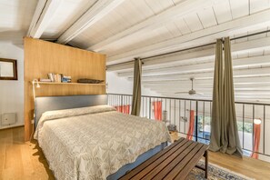 A bedroom with a bed, a wooden headboard, a shelf with books, a window with curtains, and a balcony with a railing.