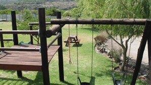 A wooden swing set, picnic table, and picnic bench in a grassy outdoor area with a view of hills.