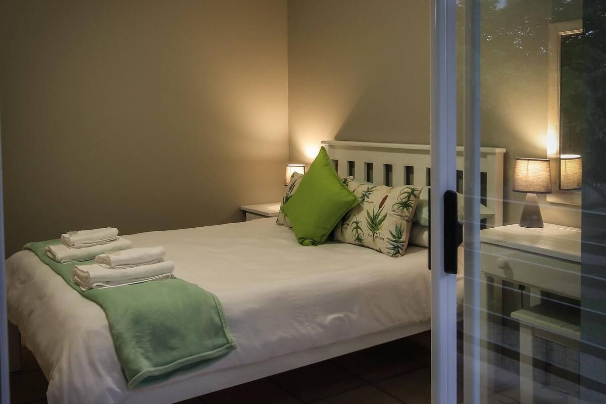 A neatly made bed with white linens and a green throw, a green pillow, and a nightstand with a lamp.