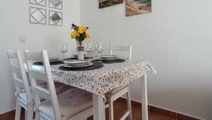Uma sala de jantar com uma mesa posta para quatro pessoas, cadeiras brancas, um candelabro e duas molduras na parede.