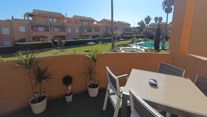 Varanda com mesa e cadeiras, plantas em vasos e vista para uma piscina e edifícios de apartamentos.