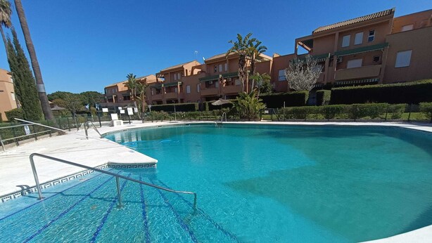 Piscina com água azul cristalina, rodeada por um deck branco e um edifício com varandas.
