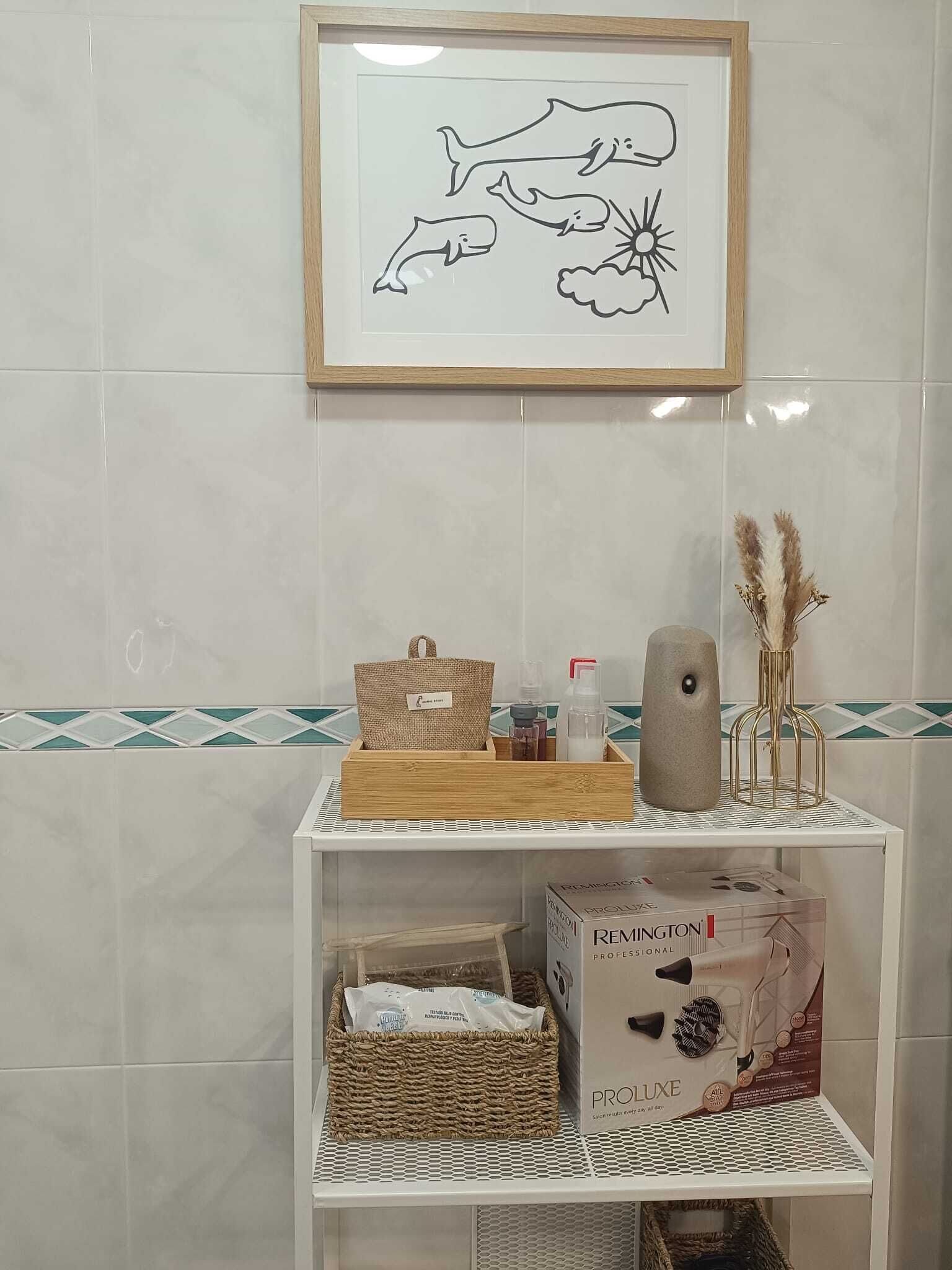 Un baño con una obra enmarcada de delfines, una estantería con artículos de tocador y una caja de secador de pelo Remington Proluxe.