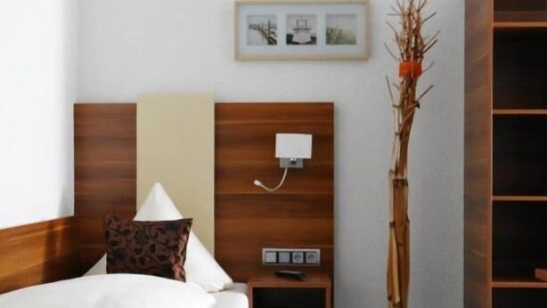 A hotel room with a bed, bedside table, lamp, wall-mounted phone, and a decorative plant. - Hotel Schützenhof mit Wellness  (Buerstadt)