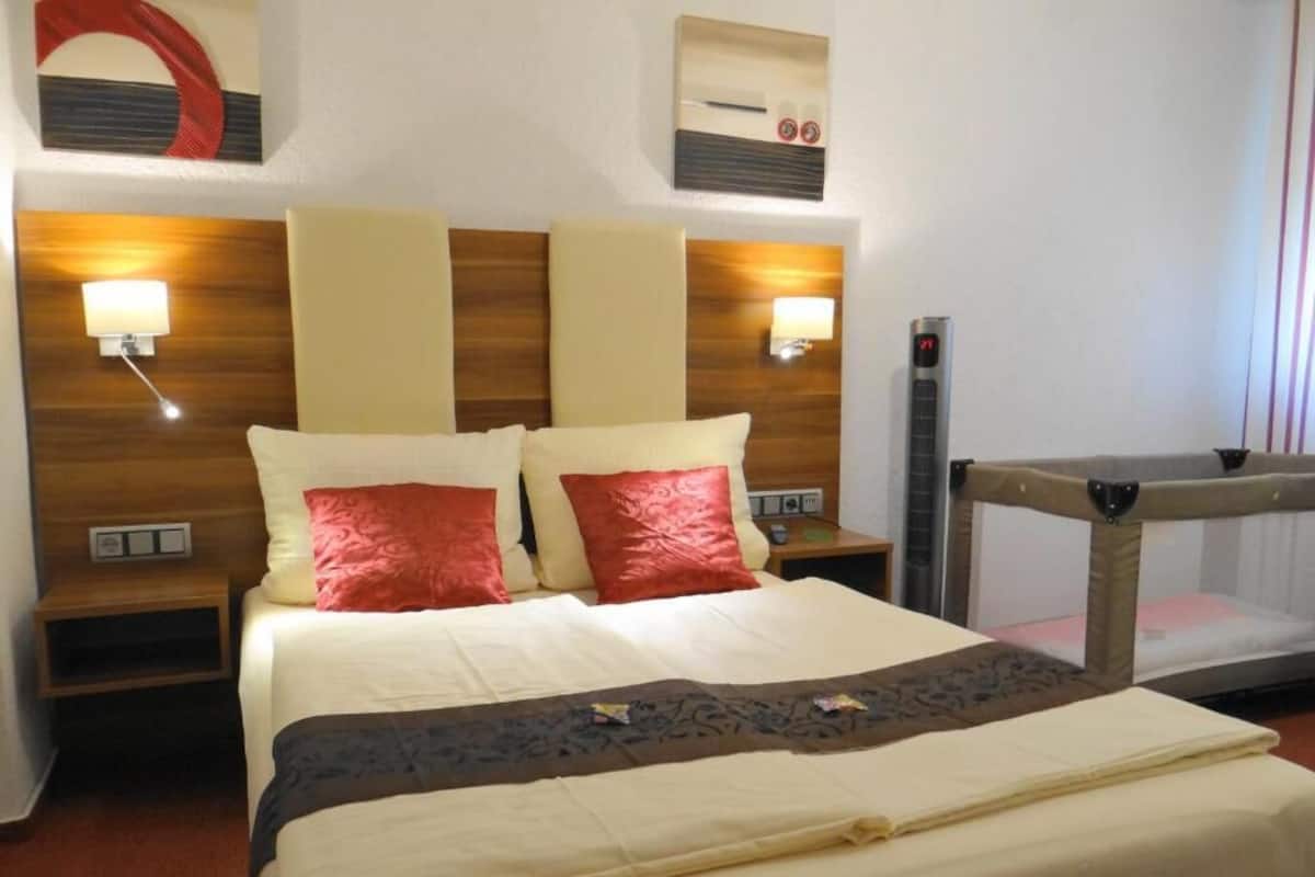 A hotel room with a large bed, two bedside tables, a nightstand, a wall-mounted heater, and a window with blinds.