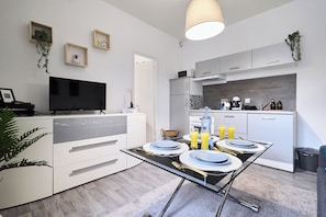 A modern kitchen with white cabinets, a dining table set with plates and glasses, a TV on a stand, and a hanging plant. - Fully equipped 4-person apartment with free public parking (Reims)