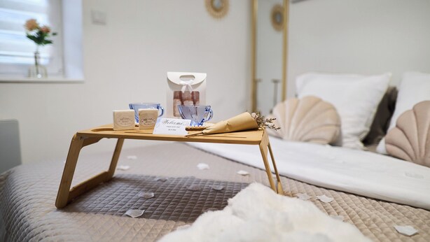 Ein ordentlich bezogenes Bett mit einer gesteppten Decke, ein Nachttisch mit Tablett und Toiletteartikeln, ein Willkommensschild und ein Fenster mit Blick nach außen.