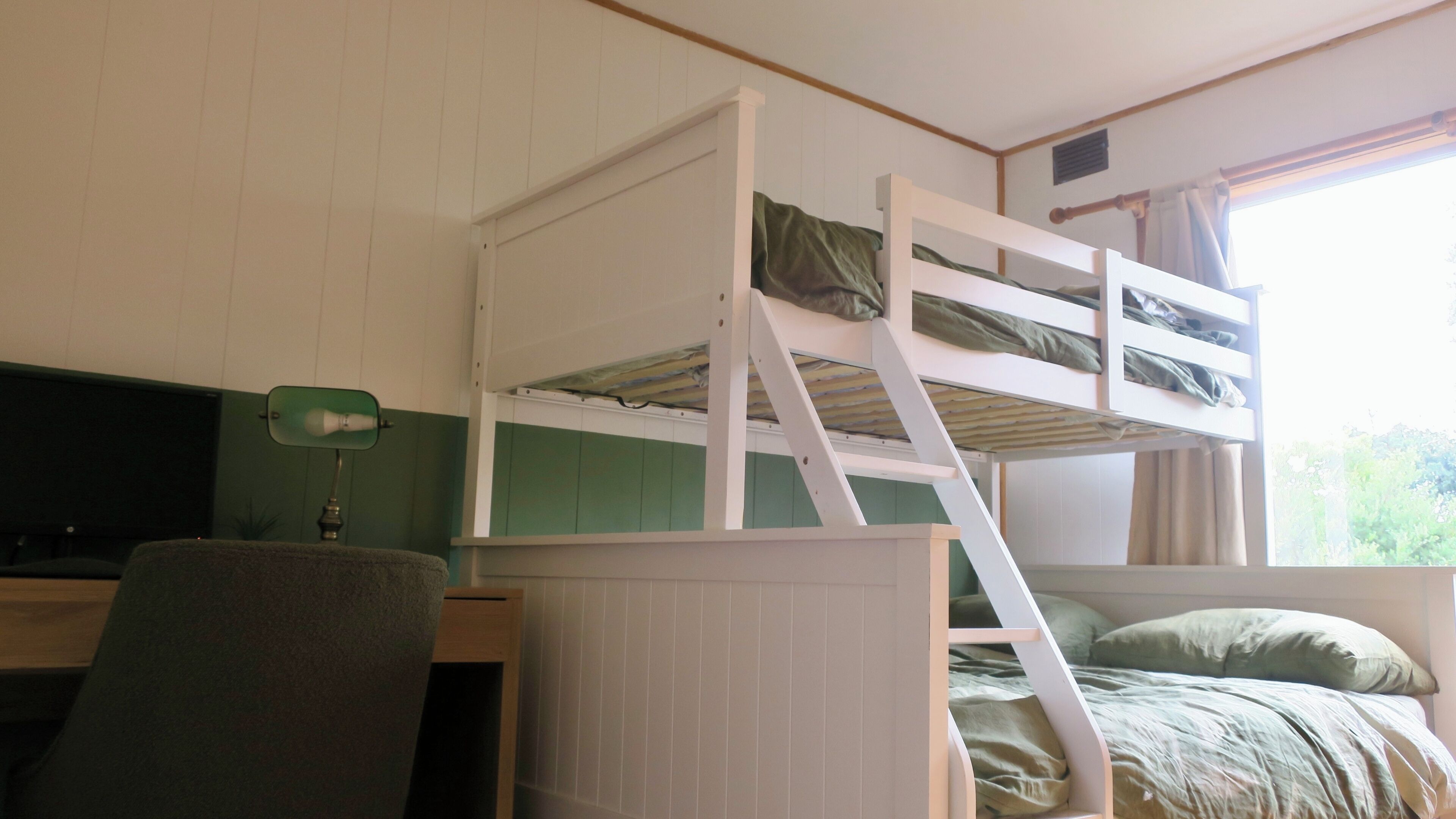 Een stapelbed met een bureau en stoel, een raam met gordijnen, en een groene wand.