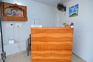 Ein Empfangsschalter mit einem Schild, auf dem 'CHECK OUT TIME 11.00 A.M.' steht, ein Wandventilator, ein Kalender und ein gerahmtes Bild einer Gottheit.