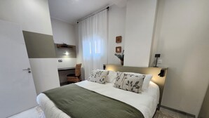 A bedroom with a bed, a desk, a chair, and a small shelf with plants.