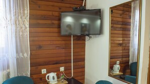 Una habitación con paneles de madera, un televisor de pantalla plana en la pared, dos sillas color turquesa, una mesita con una tetera y un control remoto, y un espejo que refleja la habitación.