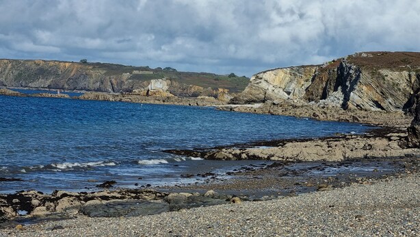 Eine Küstenlandschaft mit schroffen Klippen, einem Kiesstrand und blauem Himmel.